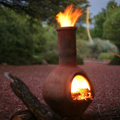 A rustic clay chiminea fire pit with a glowing fire inside and a flame extending from the top, set outdoors on a bed of stones with logs nearby.